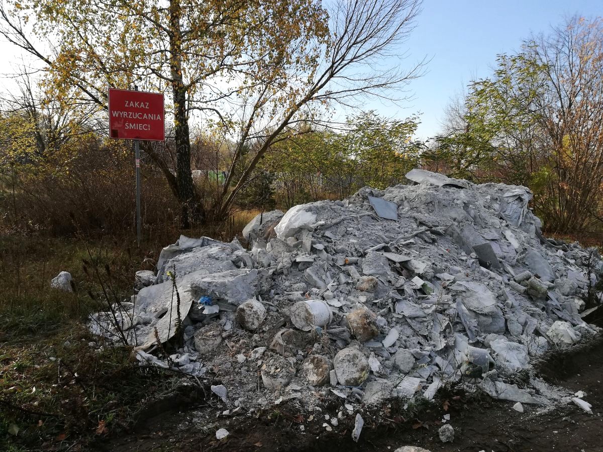 Duża sterta gruzi i innych odpadów poremontowych leży na trawie wsród drzew