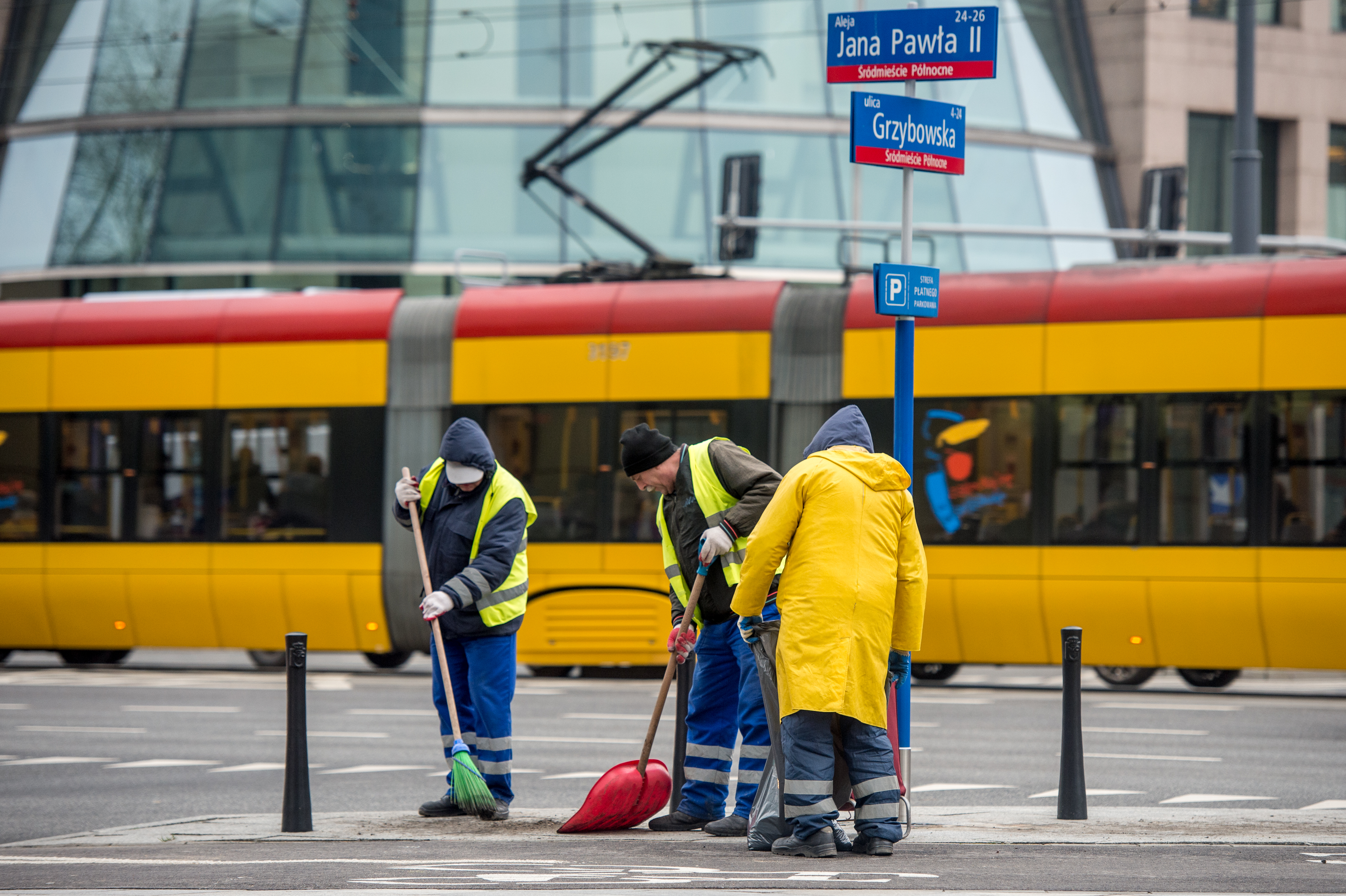 Trzej pracownicy zamiatają chodnik, jeden jest w płaszczu przeciwdeszczowym, w tle jedzie tramwaj