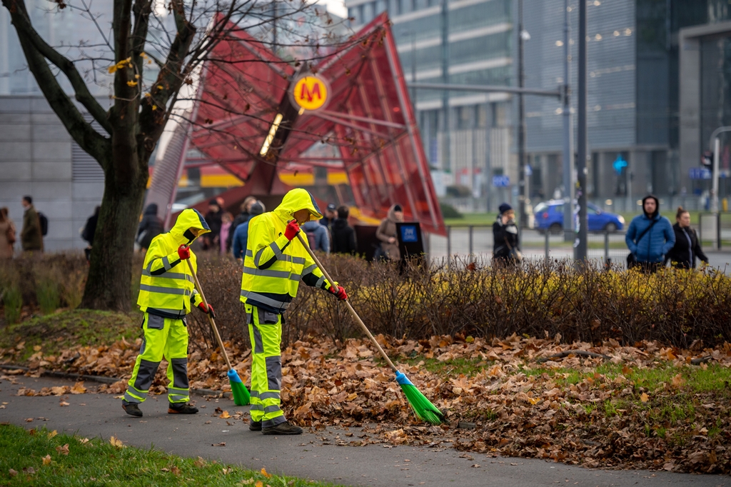 EKipa porządkowa w odblaskowych strojach zamiata jesienne liście, w tle wejście do stacji metra