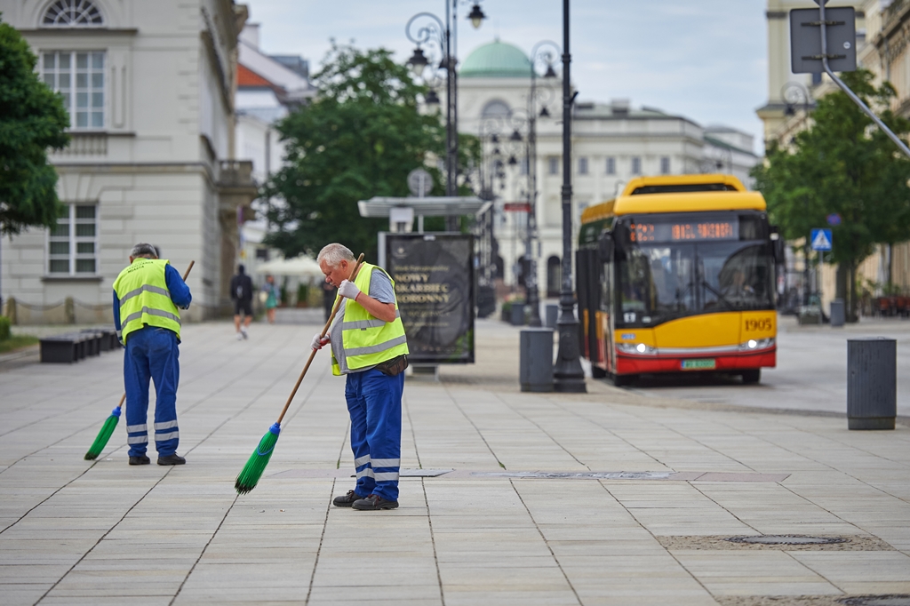 Pracownicy ekipy porządkowej zamiatają chodnik na Krakowskim Przedmieściu, w tle przystanek autobusowy, jest lato