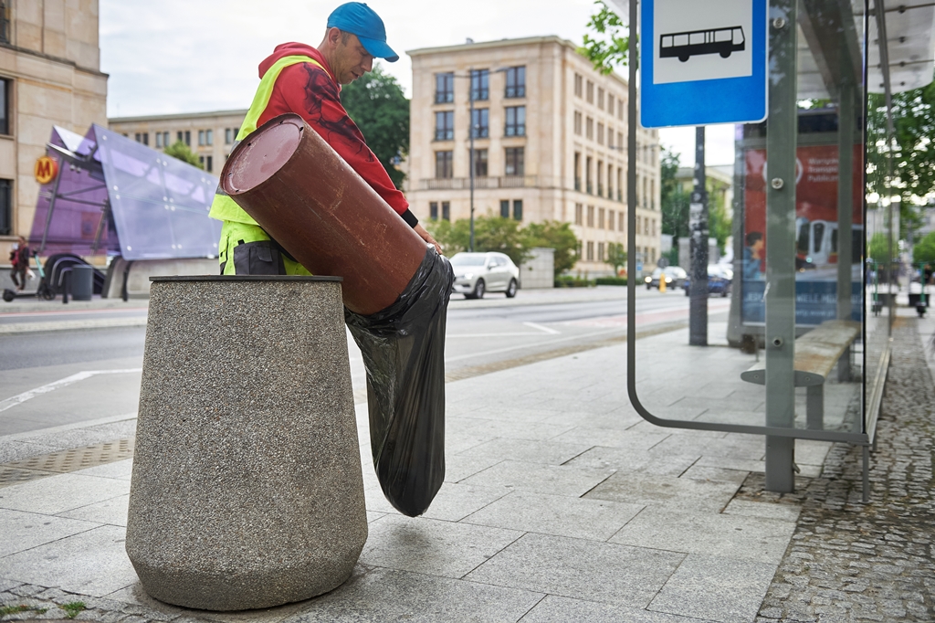Pracownik ekipy porządkowej opróżnia przystankowy kosz na śmieci, w tle wejśćie do stacji metra