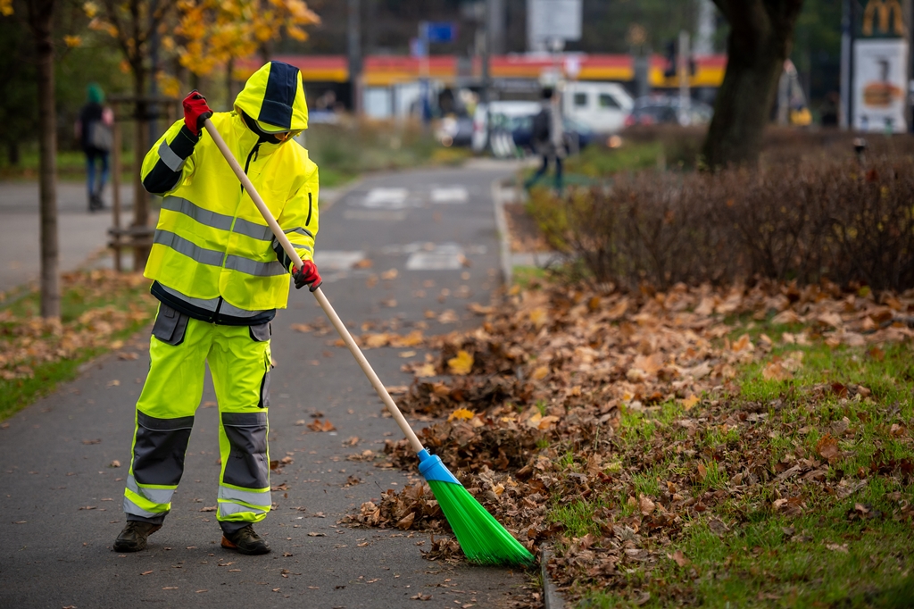 Pracownik firmy porząkowej w odblaskowym stroju zamiata kolorowe liscie z trasy dla rowerów