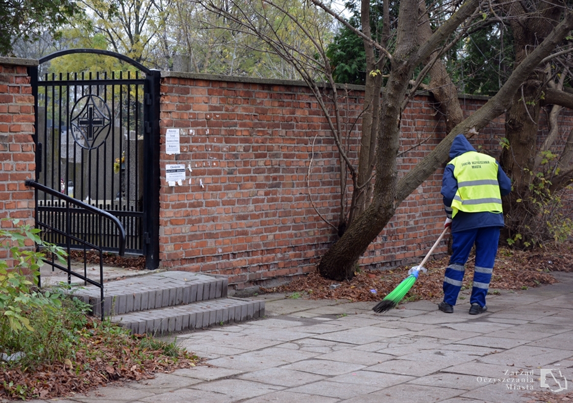 Pracownik ekipy sprzątającej zamiata liście z chodnika przed bramą cmentarza