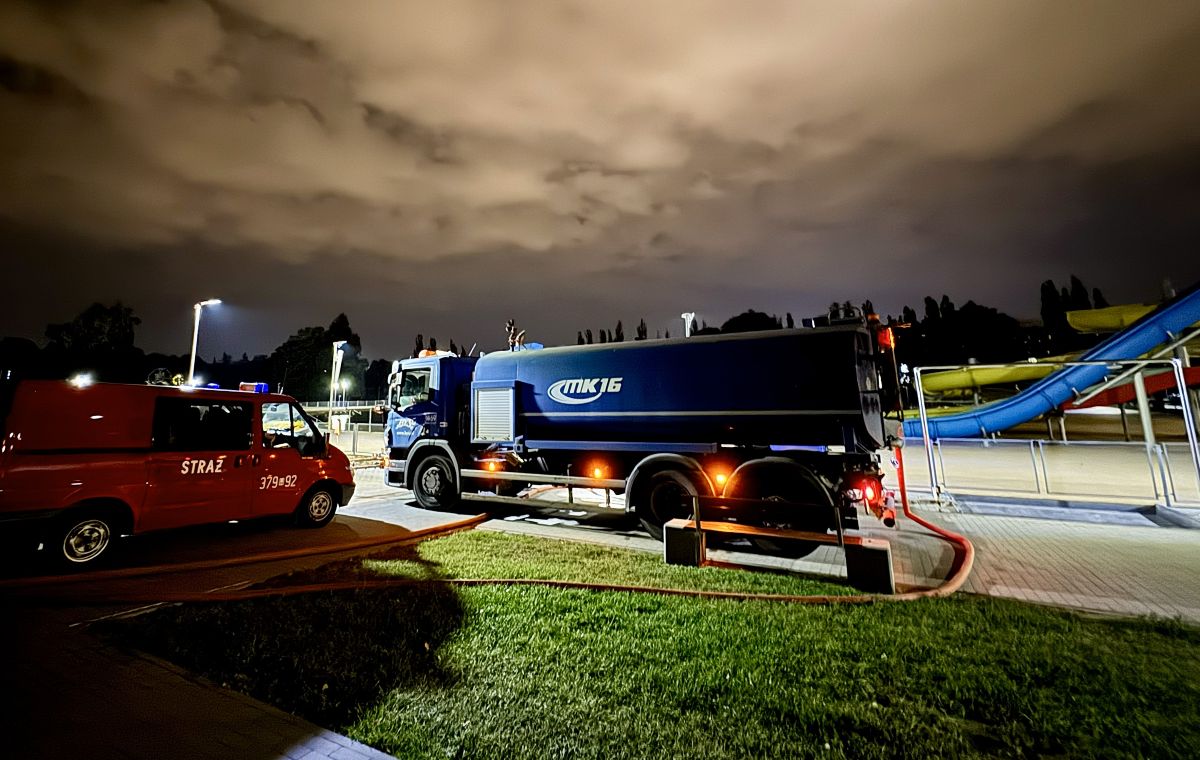 Duży pojazd - zmywarka pobiera wodę z odkrytego basenu. Jest noc, z prawej strony widać samochód straży pozarnej, z lewej - zdjeżdżalnie do basenu