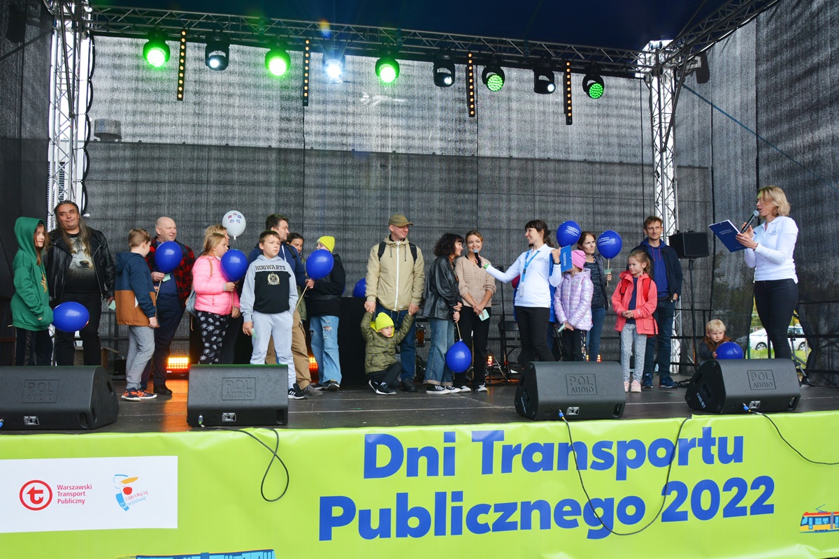 Konkurs - na scenie stoją rodzice i dzieci, jedna prowadząca czyta do mikrofonu pytania, druga prowadząca podsuwa jednej z par mikrofon do odpowiedzi, na dole banner "Dni Transportu Publicznego 2022"