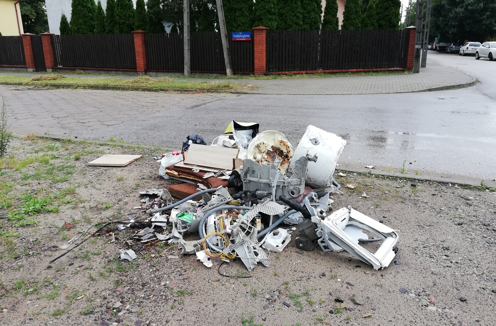 Sterta śmieci leży na terenie zieleni przy ulicy, wśród śmieci fragmenty sprzętów AGD, elementy z mebli