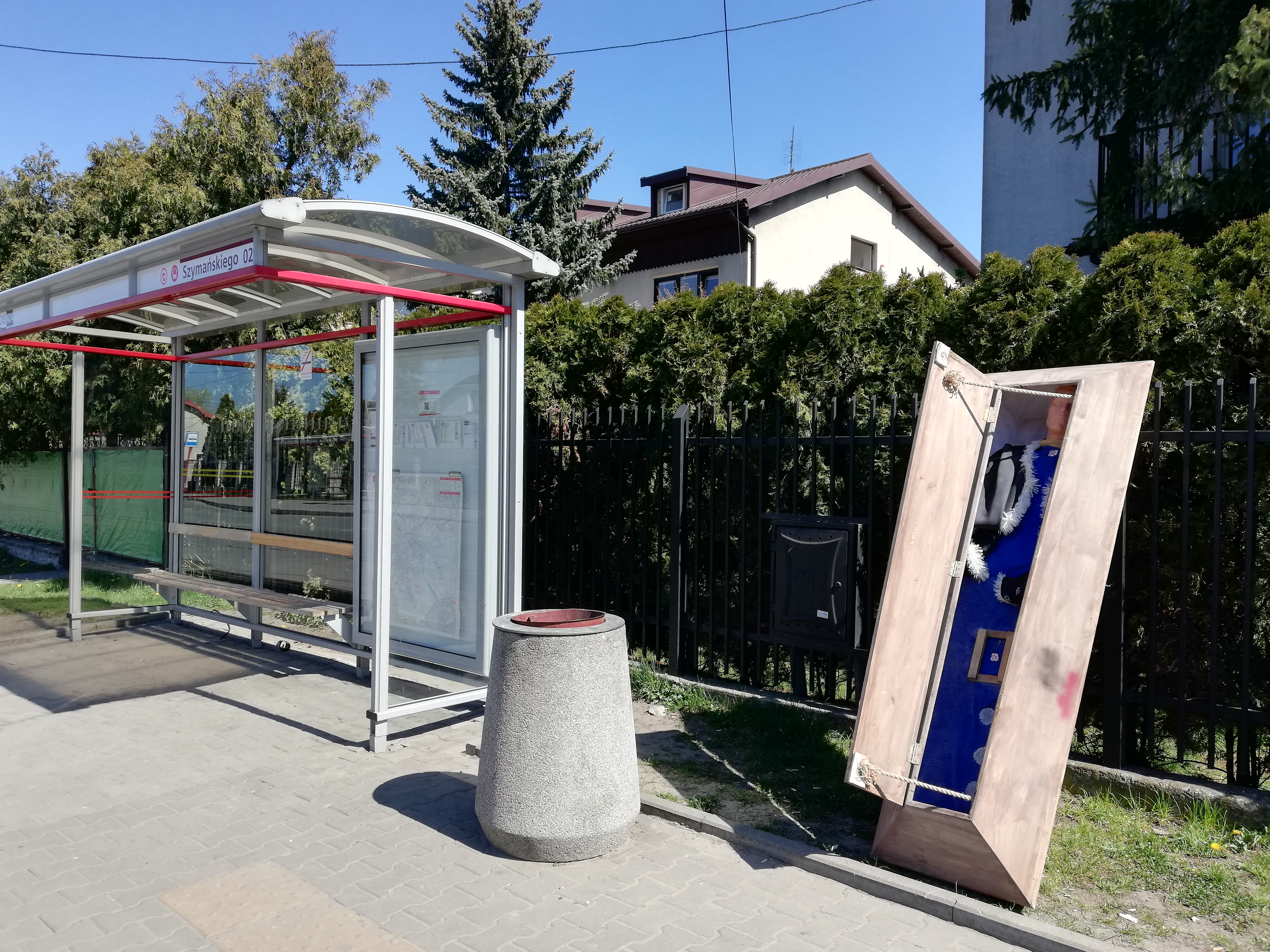 Przystanek autobusowy, obok stoi drewniana trumna z manekinem w środku