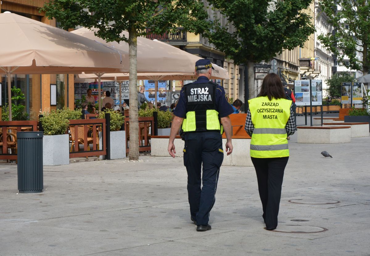 Wspólny patrol kontrolerki ZOM i strażnika miejskiego idzie przez pl. Pięciu Rogów w Warszawie, w tle drzewa i ogróddek kawiarniany