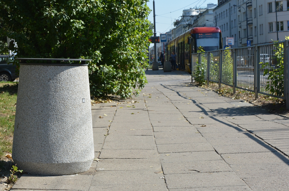 Okrągły betonowy kosz na śmieci z żeliwną pokrywą stoi na chodniku, z prawej strony torowisko tramwajowe