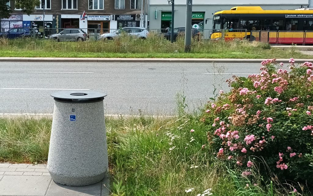 Betonowy kosz na śmieci z pokrywą stoi na chodniku, obok trawnika. Na koszu jest plakietka z oznaczeniem budżetu obywatelskiego. W tle ulica, którą jedzei autobus.