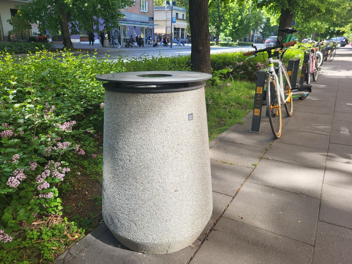 Na pierwszym planie betonowy kosz na śmieci z metalową pokrywą, stoi na chodniku obok żywopłotu. W tle rowery zaparkowane w stojakach.
