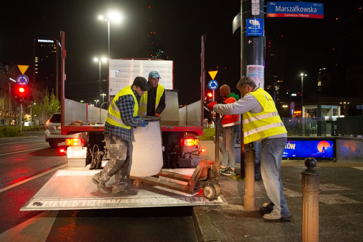 Noc, chodnik w mieście, ekipa porządkowa w odblaskowych kamizelkach za pomocą specjalnego wózka zdejmuje betonowy kosz na śmieci z paki samochodu, gdzie stoją kolejne kosze