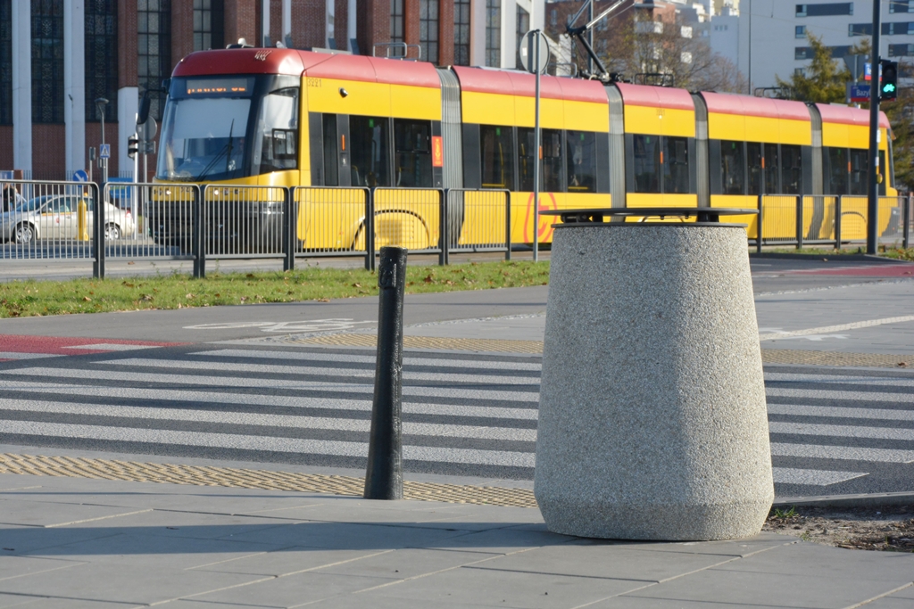 Betonowy kosz na śmieci stoi przy przejściu dla pieszych, w tle tramwaj