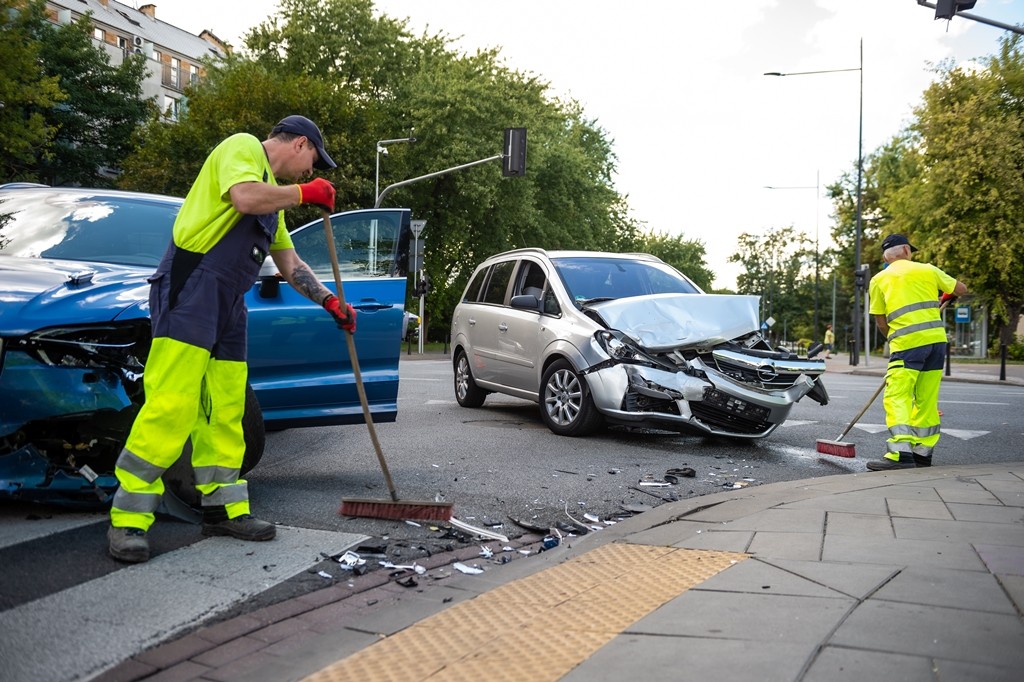 Dwaj pracownicy Pogotowia Porządkowego zamiatają jezdnię z fragmentów karoserii, na drugim planie dwa rozbite samochody po wypadku