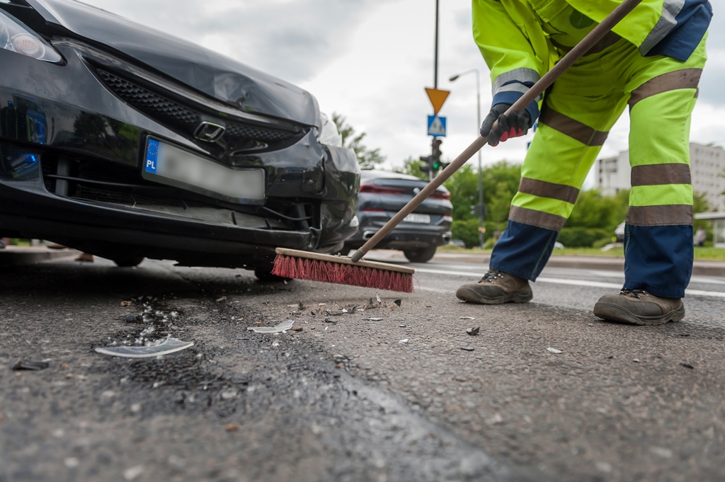 Przód rozbitego samochodu i człowiek, który zamiata z jezdni fragmenty karoserii, ujęcie od ramion w dół