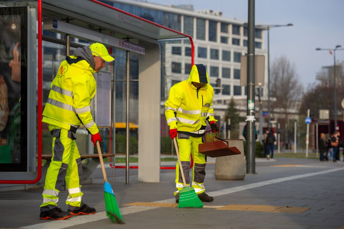 Dwóch pracowników firmy porządkowej w odblaskowych strojach roboczych zamiata chodnik na przystanku