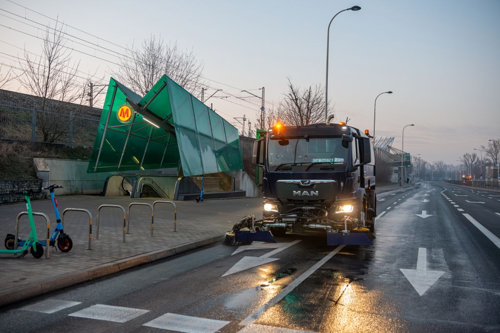 Zamiatarka jedzie po ulicy, z lewej strony wejście do stacji metra