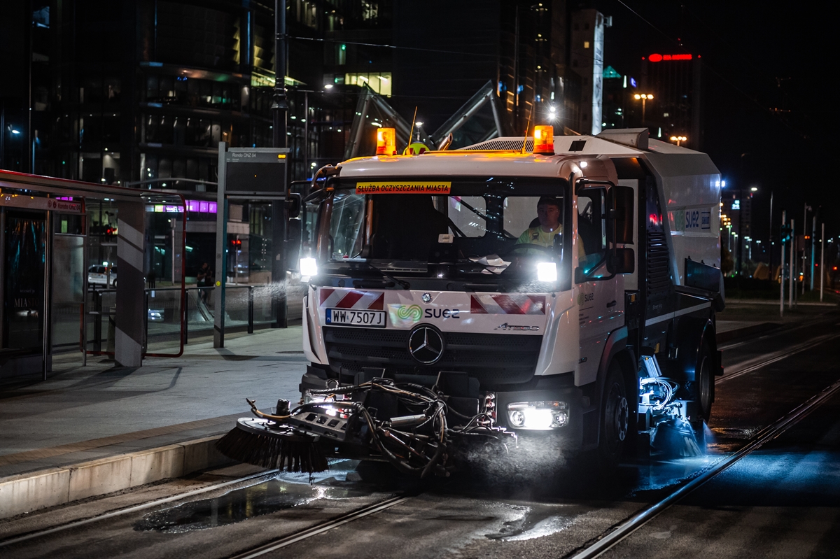 Zamiatarka nocą czyści torowisko tramwajowe, przejeżdża obok przystanku
