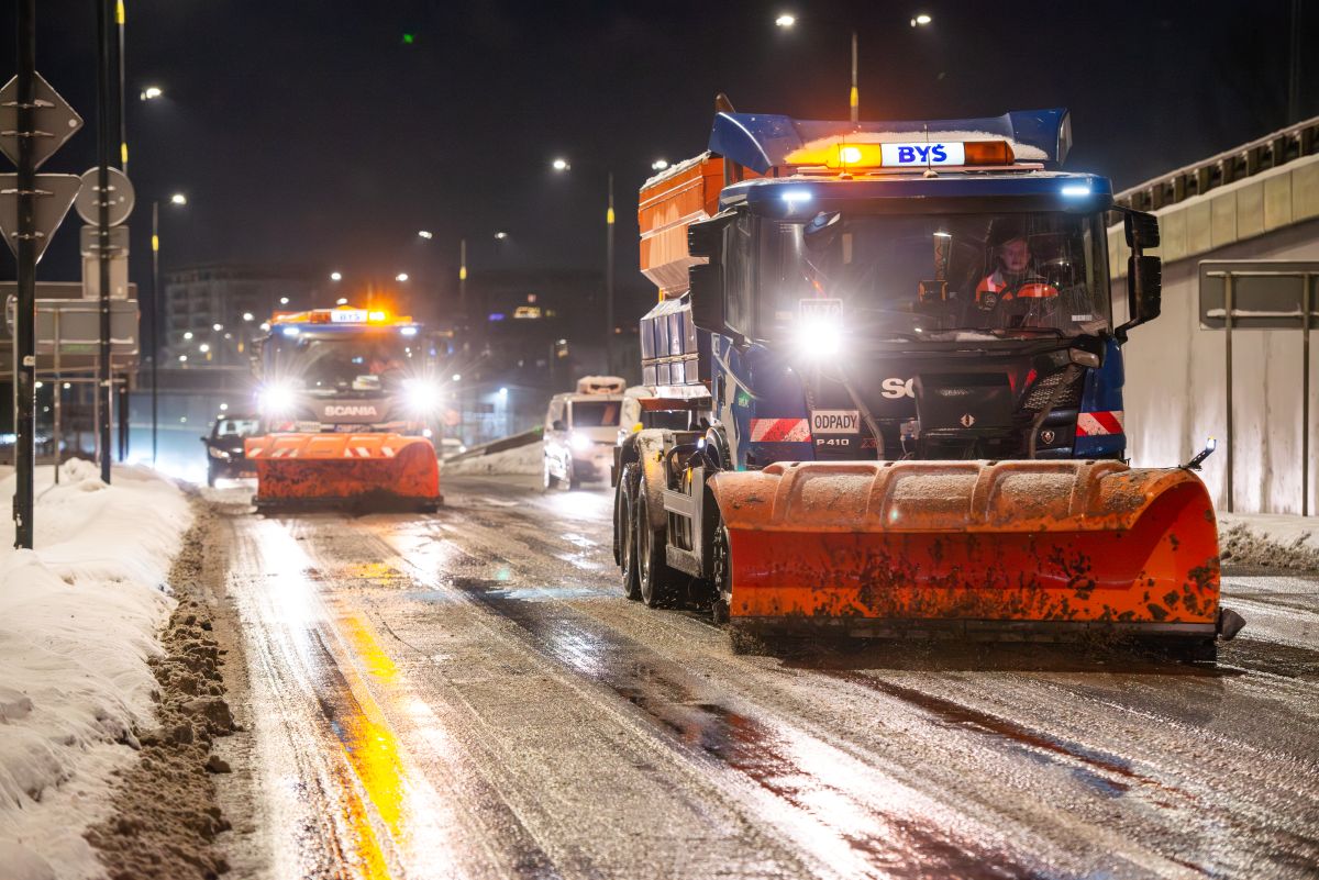 Dwie posypywarki działają na ulicy, jest noc, z lewej strony ulicy leży zgarnięty śnieg