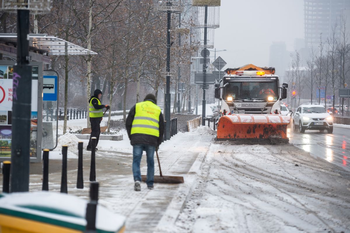 Mioasto zimą, na ulicy pracuje pług, który odgarnia śnieg, na chodniku po lewej stronie pracownicy z szuflami sprzątają śnieg z przystanku autobusowego
