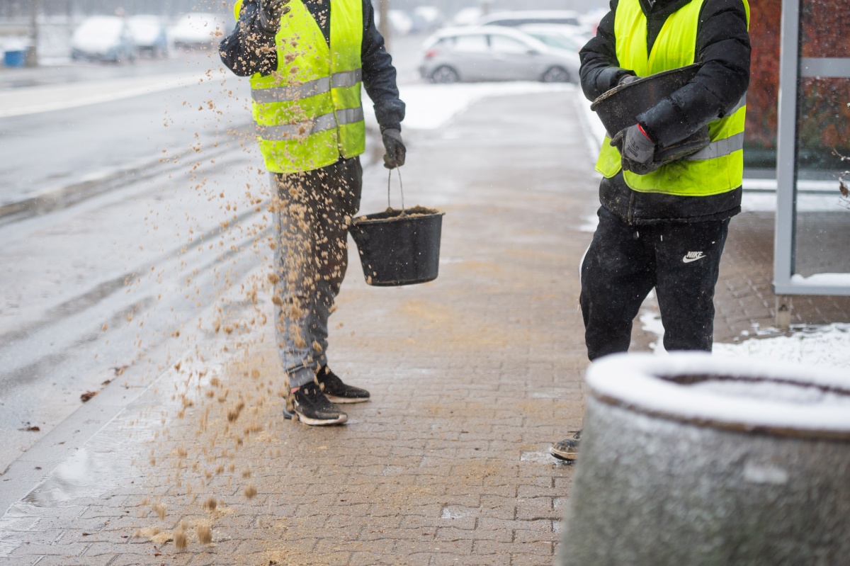 Dwóch pracowników firmy porządkowej na przystanku autobusowym posypuje chodnik piaskiem. Jest zima, wokoło widać śnieg.