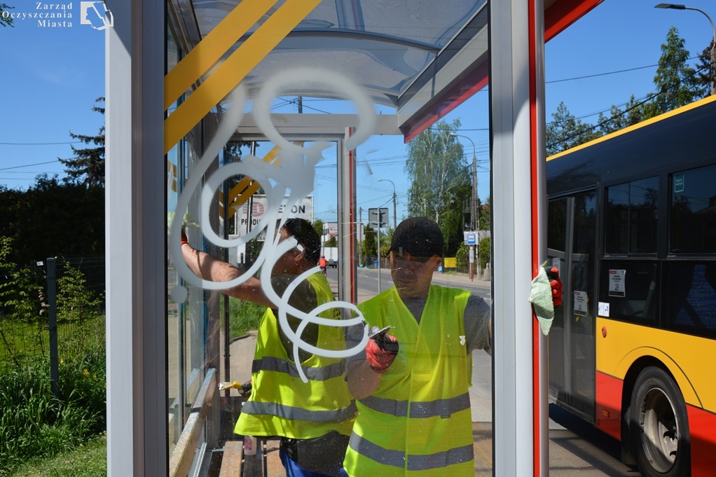 Pracownicy ZOM w kamizelkach odblaskowych usuwają pseudograffiti z szyby wiaty przystankowej. Po prawej stronie widać autobus, który zatrzymał się na przystank. W lewym górnym rogu znajduje się logo Zarządu Oczyszczania Miasta.