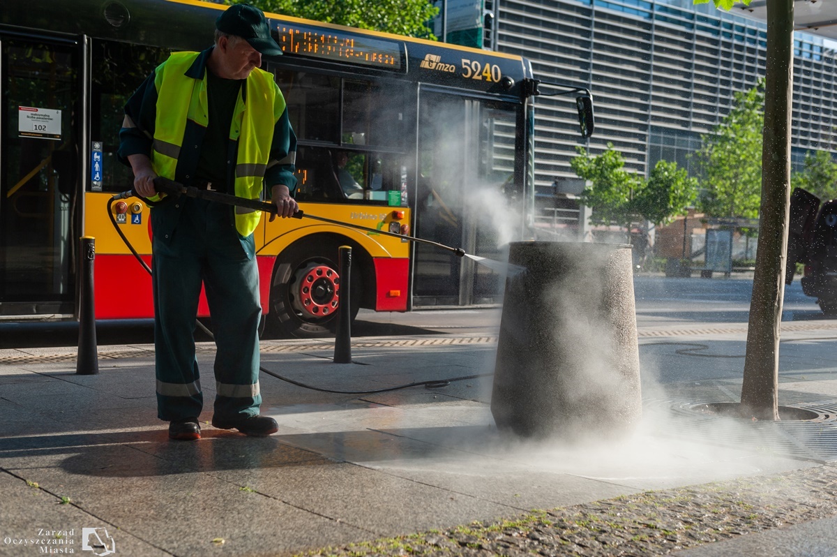 Pracownik firmy porządkowej myje kosz myjką ciśnieniową, w tle widać autobus miejski