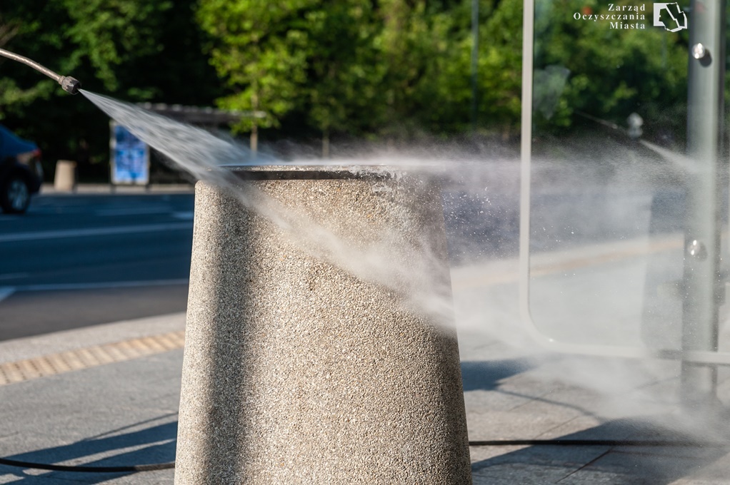 Zbliżenie na betonowy kosz uliczny podczas mycia wodą pod ciśnieniem. Z lewej strony widać dyszę myjki. W prawym górnym rogu znajduje się logo Zarządu Oczyszczania Miasta.