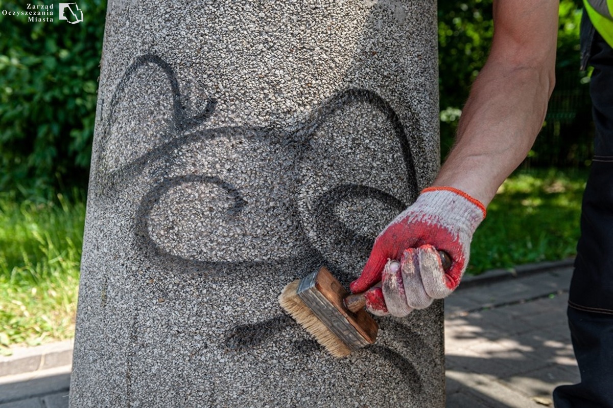Zbliżenie na betonowy kosz na którym jest wysprejowany napis. Widać rękę pracownika trzymającego pędzel w biało-czerwonej rękawiczce. Pędzlem nakłada na kosz bezbarwny preparat czyszczacy.