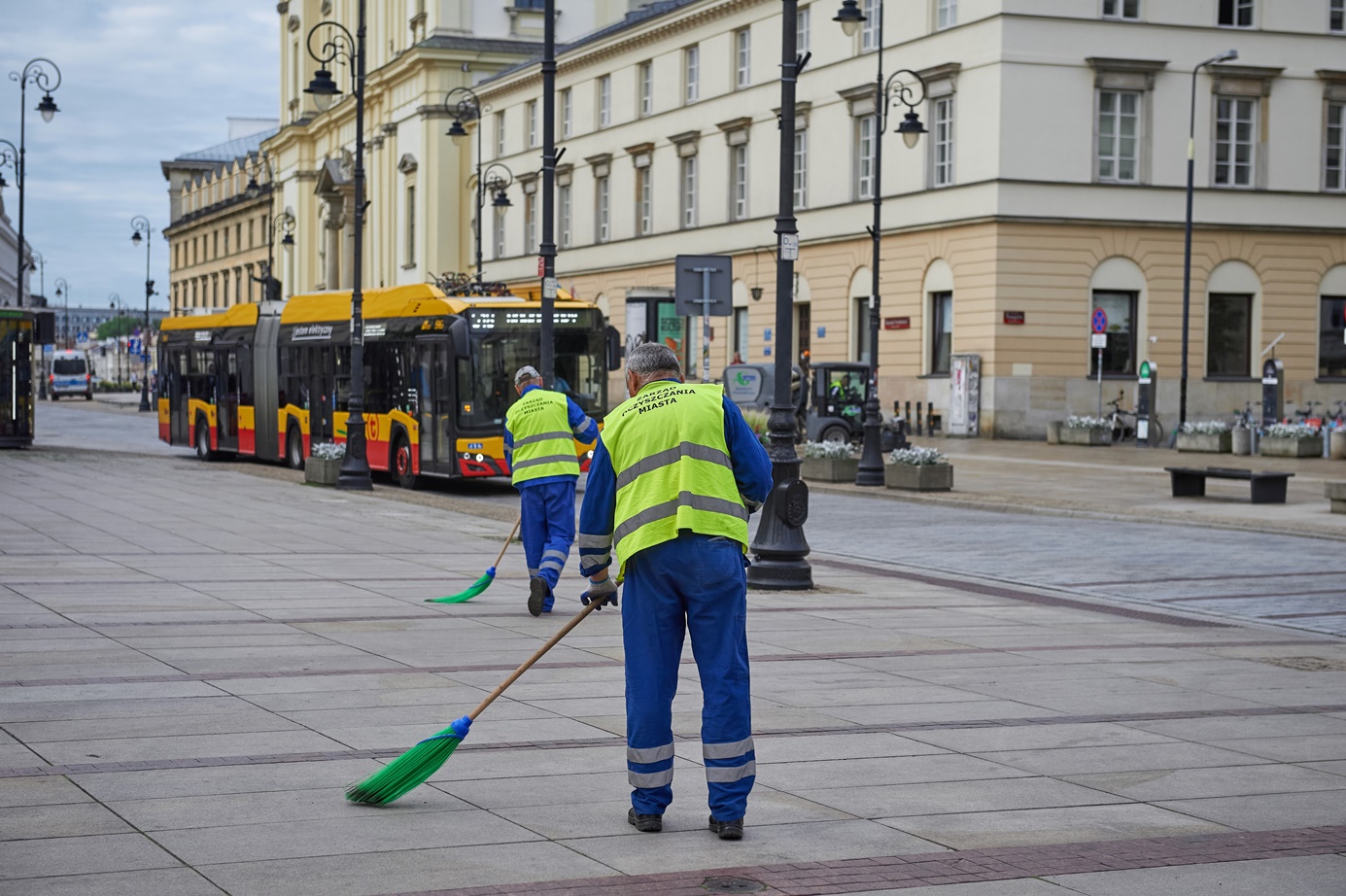 Pracownicy służby oczyszczania miasta zamiatają chodnik na Krakowskim Przedmieściu, w tle jedzie autobus.