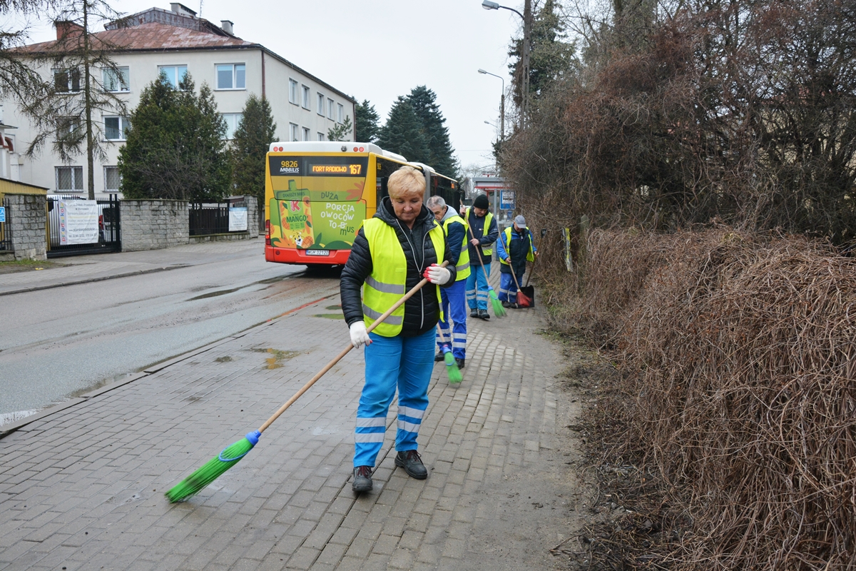 Na pierwszym planie panie Ewa Groszek w stroju roboczym, z miotłą, na drum planie dwóch pracowników zamiata miotłami chodnik, z tyłu przystanek autobusowy