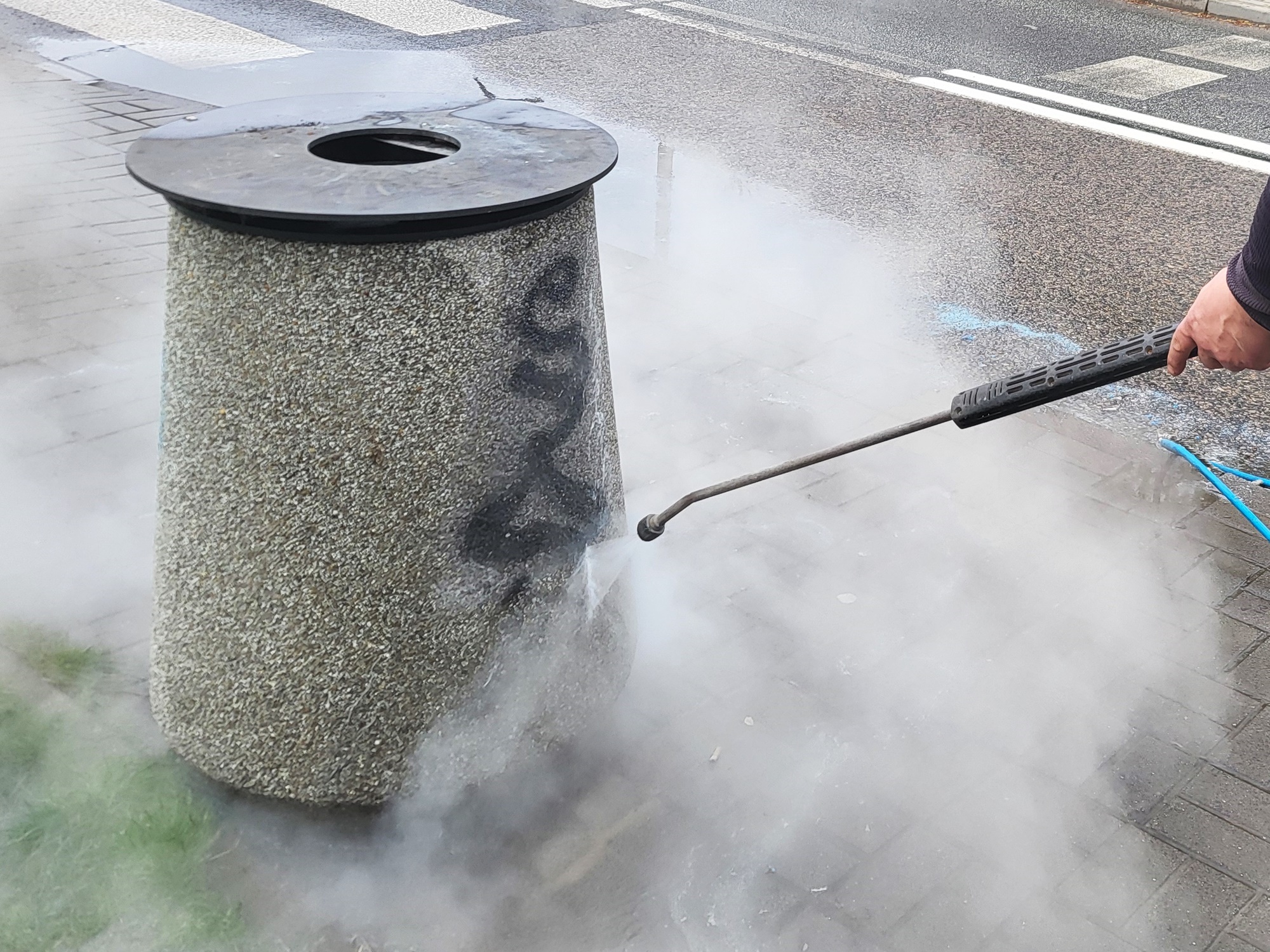 Pomazany betonowy kosz na chodniku, widać myjkę, z której woda pod ciśnieniem czyści powierzchnię kosza