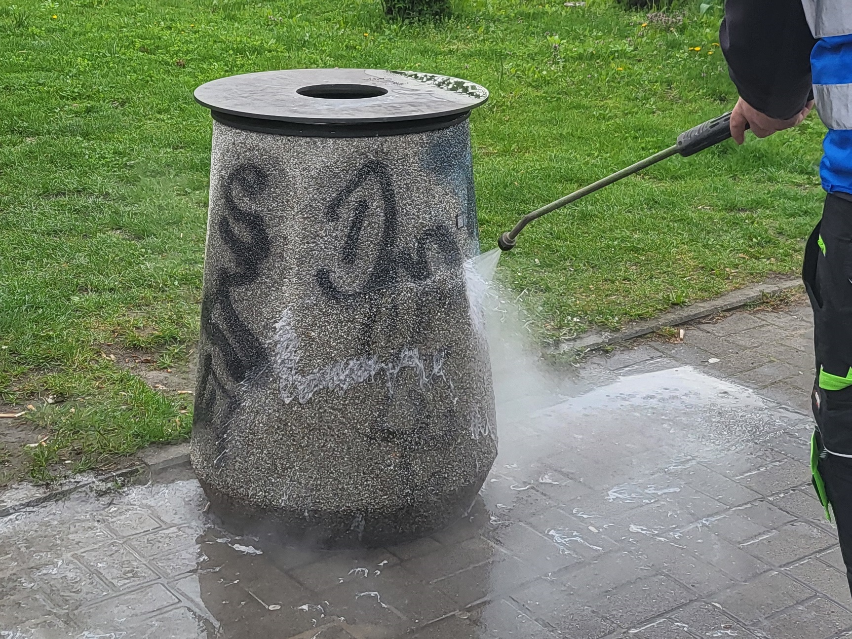 Pomazany betonowy kosz na chodniku, pracownik firmy porządkowej myje go z użyciem myjki cośnieniowej