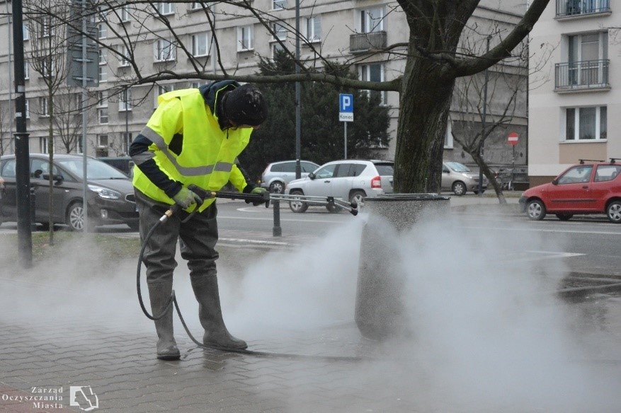 Pracownik firmy porządkowej myje wodą pod ciśnieniem betonowy kosz