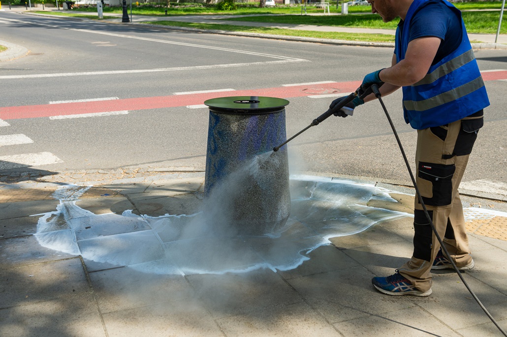 Kosz betonowy stoi na chodniku, pracownik firmy porządkowej usuwa pseudograffiti wodą pod ciśnieniem. W tle ścieżka rowerowa i ulica