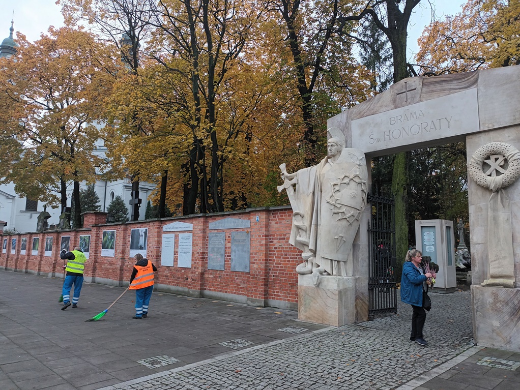 Ekipa porządkowa zamiata chodnik przy cmentarzu, w tle mur nekropolii oraz brama wjazdowa.