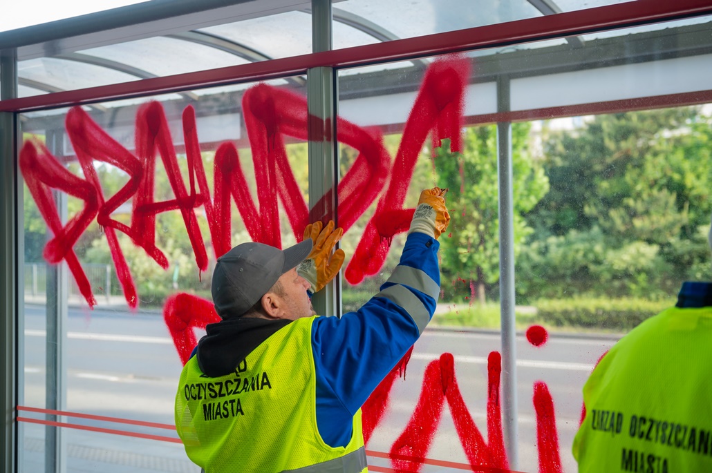 Ekipa porządkowa usuwa pseudograffiti z wiaty przystankowej, w tle ulica i drzewa