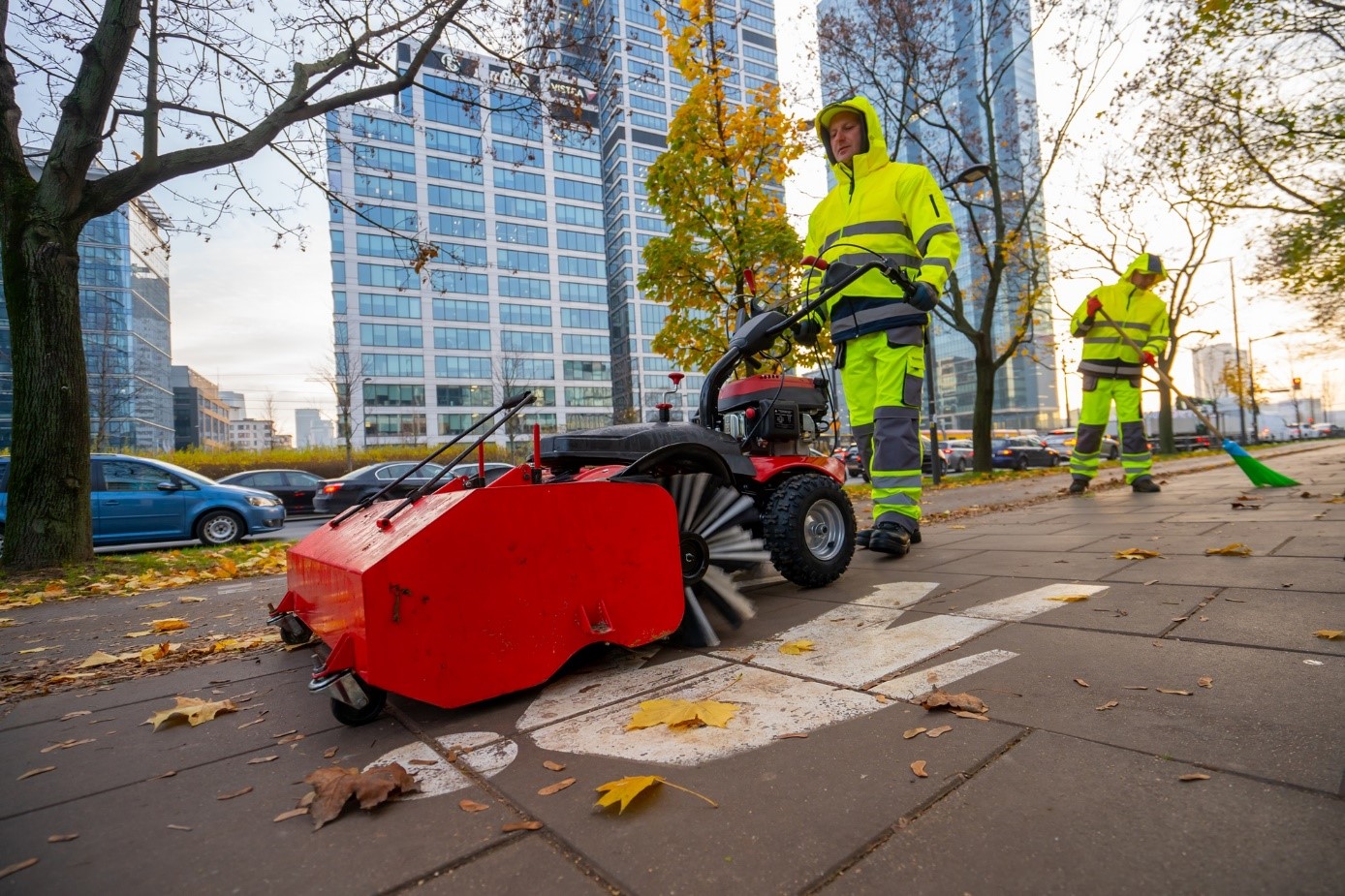 Pracownik formy porządkowej zamiata chodnik z liści przy użyciu mechanicznej zamiatarki. W tle drzewa, ulica i biurowiec