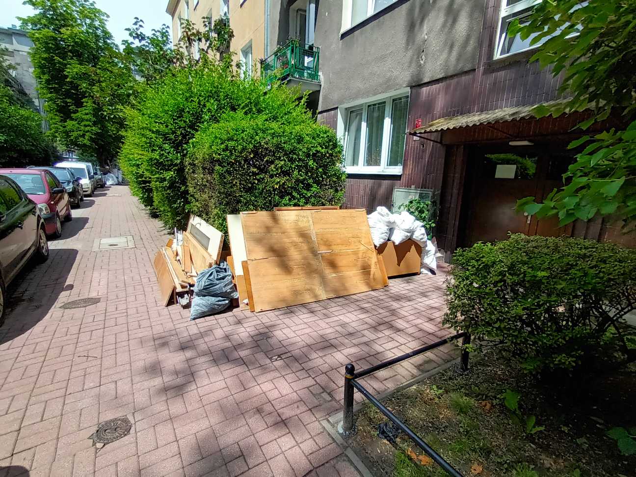 Stare meble pozostawione na chodniku przed wejście do bloku mieszkalnego, w tle żywopłot i zaparkowane auta