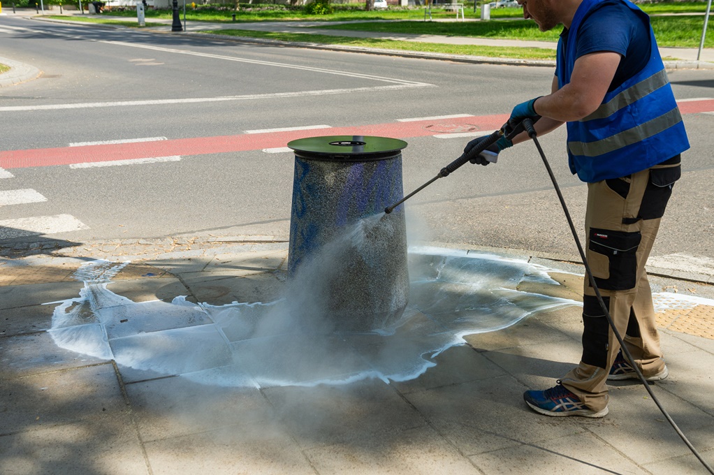 Mężczyzna w kamizelce odblaskowej myje myjką ciśnieniową pomazany kosz. Kosz stoi na chodniku, w tle ulica i ścieżka rowerowa