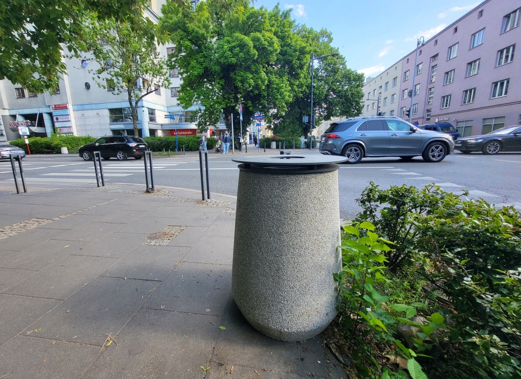 Betonowy kosz stoi na chodniku przy przejściu dla pieszych, w tle ulica i bloki mieszkalne