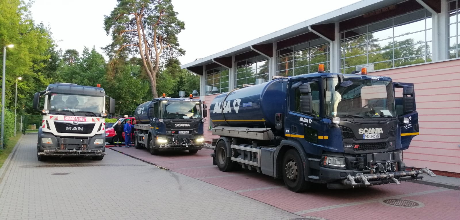 Zmywarki stojąf na parkingu przed basenem, pobierają wodę z pływalni, w tle budynek i drzewa