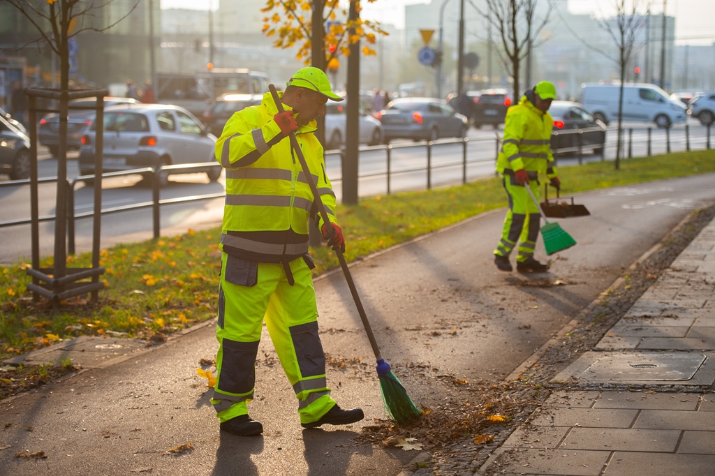 Dwóch mężczyzn w strojach roboczych zamiata ścieżkę rowerową, w tle ulica