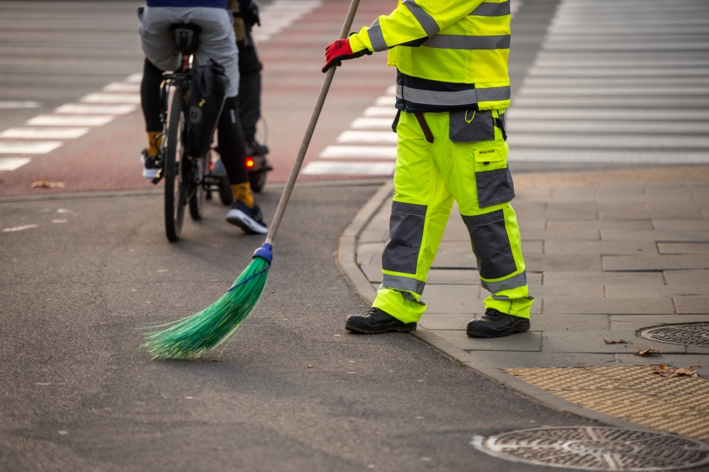 Pracownik ekipy porządkowej zamiata drogę rowerową, w tle osoba na rowerze i osoba na hulajnodze