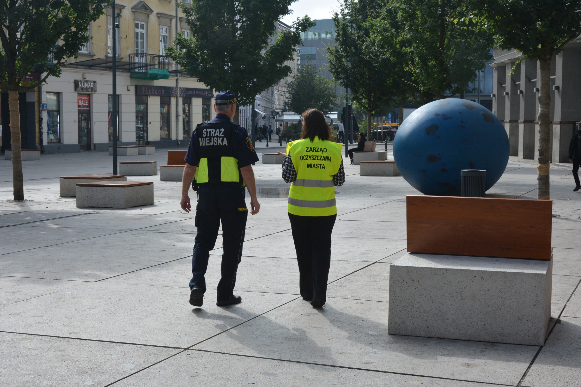 Funkcjonariusz Straży Miejskiej i pracownica Zarządu Oczyszczania Miasta sprawdzają miasto pod kątem czystości. Patrolują plac, po prawej stronie jest rzeźba wielkiego niebieskiego jajka